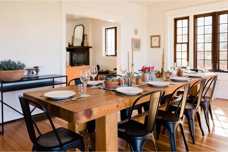 Mesa de comedor de madera puesta para una comida, con platos blancos, copas de vino, candelabros dorados con velas, macetas de suculentas y sillas metálicas negras, en un comedor luminoso.