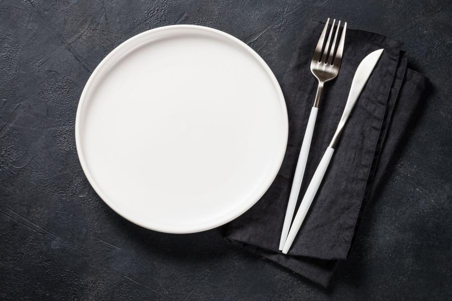 Vista cenital de un plato blanco mate junto a una servilleta de lino negra doblada con un tenedor y un cuchillo de mango blanco encima, sobre una superficie oscura de textura pizarra.