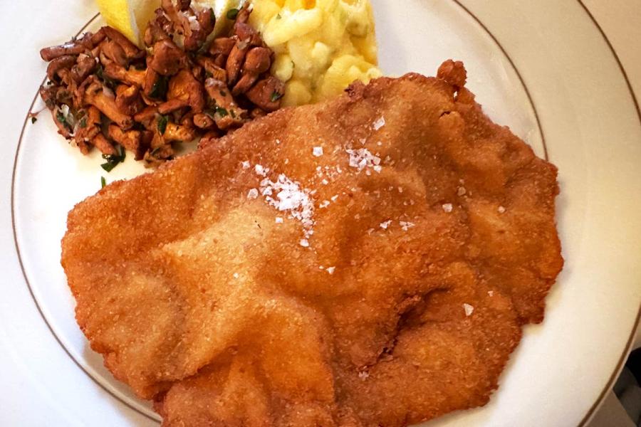 Schnitzel dorado con sal en escamas, acompañado de ensalada de patata y setas salteadas en un plato blanco.