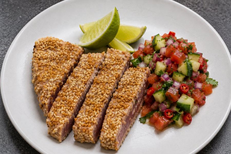 Lomos de atún sellado con costra de sésamo, dorado por fuera y rosado por dentro, cortado en tiras, servido con gajos de lima y una salsa fresca de tomate, pepino y cebolla roja.