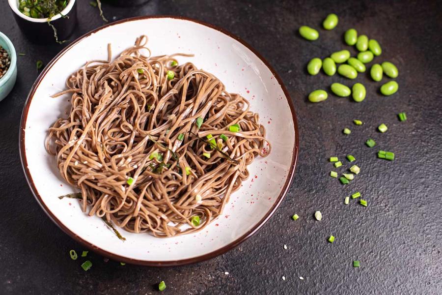 Cuenco blanco con fideos soba fríos aliñados, espolvoreados con cebolleta picada y hebras de alga, junto a granos de edamame y sésamo esparcidos sobre una superficie oscura.