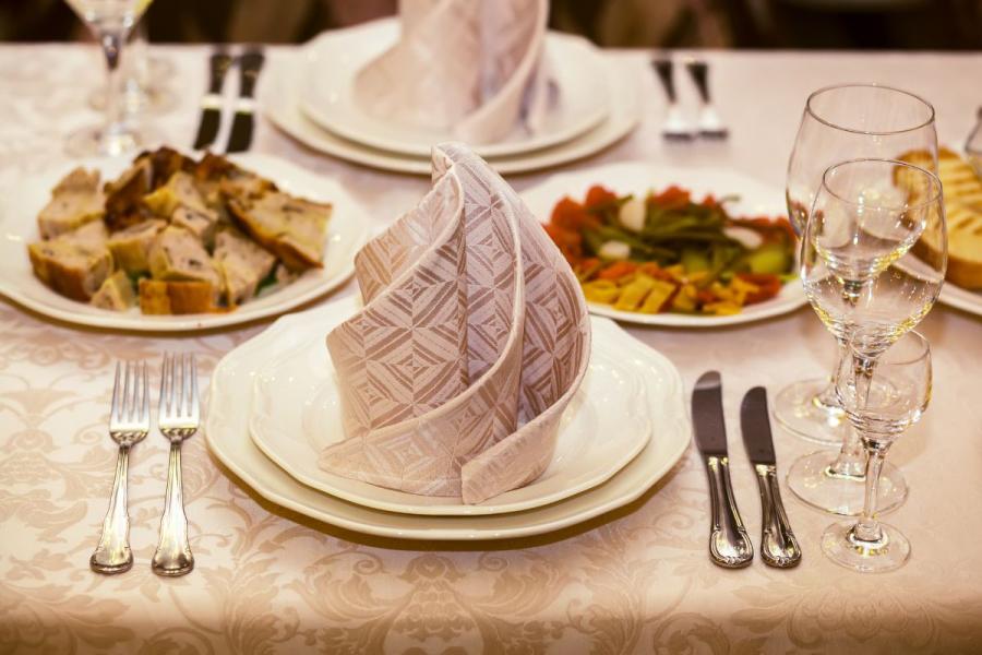 Mesa de restaurante elegante con mantel de damasco, servilletas de tela dobladas en pie, copas de vino de cristal y varios platos de comida servidos al fondo.