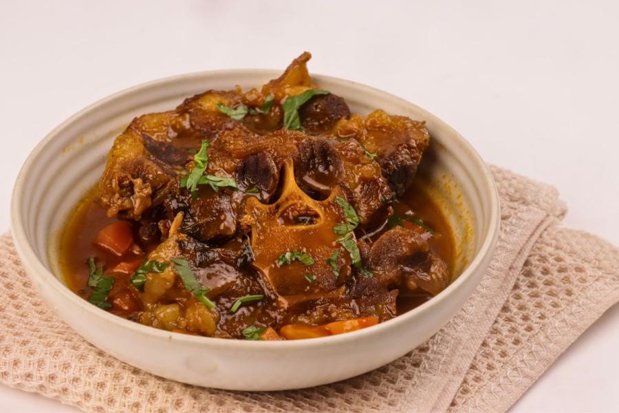 Cuenco de cerámica clara con rabo de toro braseado en una salsa espesa y brillante, con trozos de zanahoria y perejil picado por encima, sobre un paño de cocina beige.