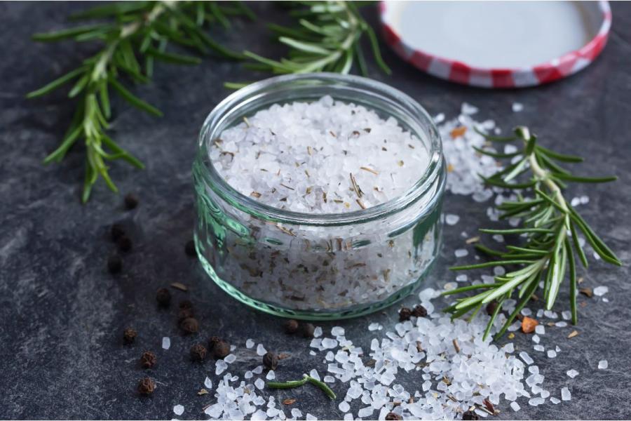 Tarro de cristal con sal gruesa aromatizada con romero, rodeado de ramitas de romero fresco y granos de pimienta negra, sobre una superficie oscura de pizarra.