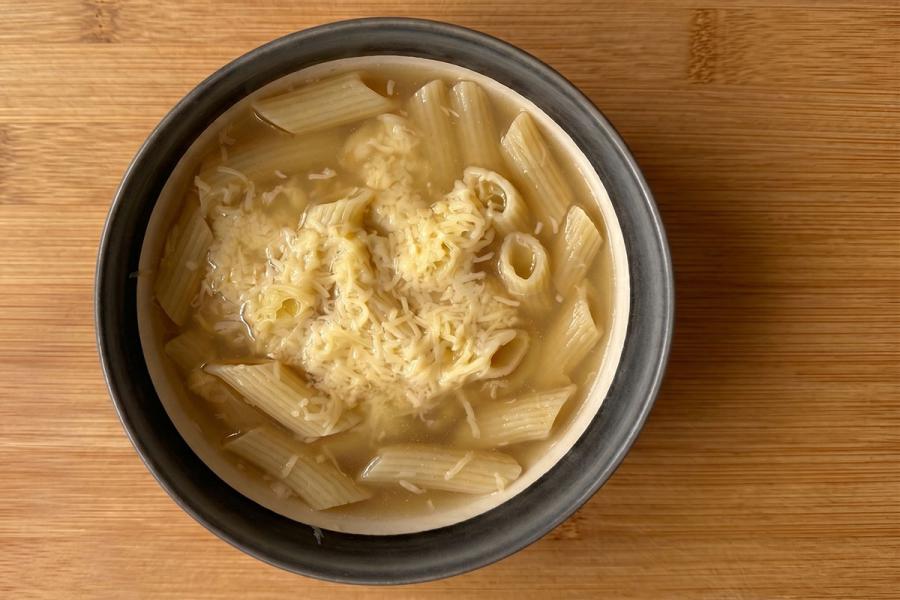 Cuenco de sopa de macarrones con queso rallado por encima, servido en un bol de cerámica gris sobre una mesa de madera.