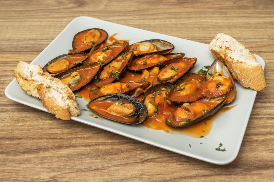 Mejillones en escabeche con salsa de tomate y hierbas, servidos en un plato blanco junto a rebanadas de pan tostado.