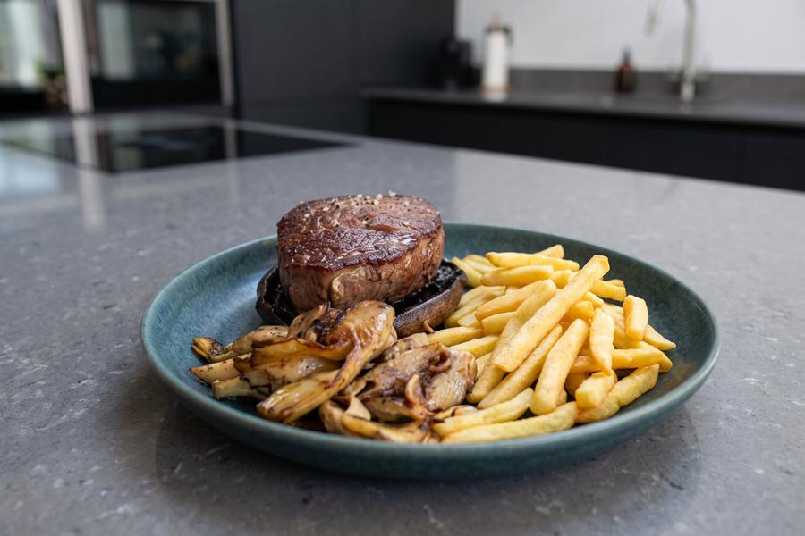 Sobre la encimera de la cocina, un plato con un tournedós sobre una cabeza de seta Porotbello acompañado de aclcachofas y patatas paja.