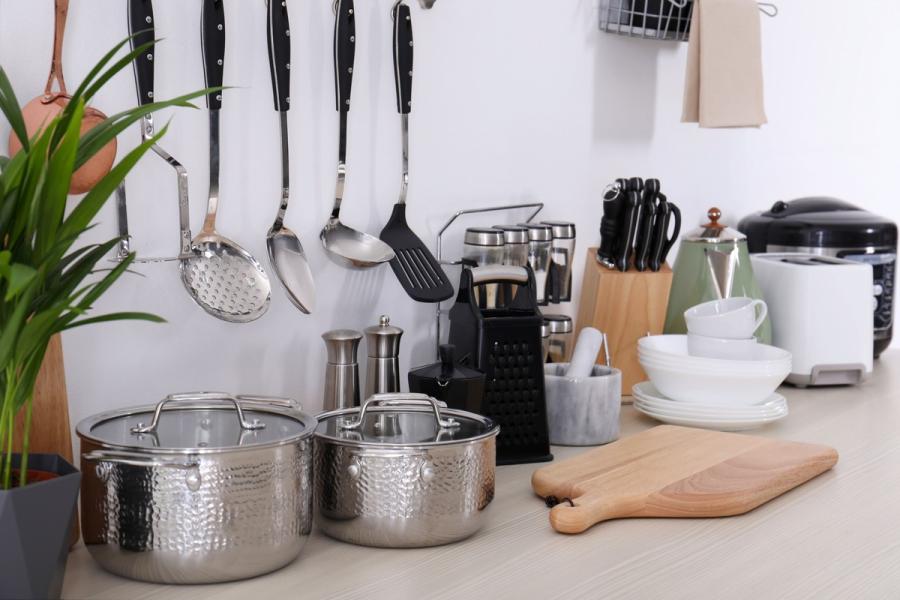 Encimera de cocina con utensilios de acero inoxidable colgados, dos ollas, tabla de madera, vajilla blanca y pequeños electrodomésticos.