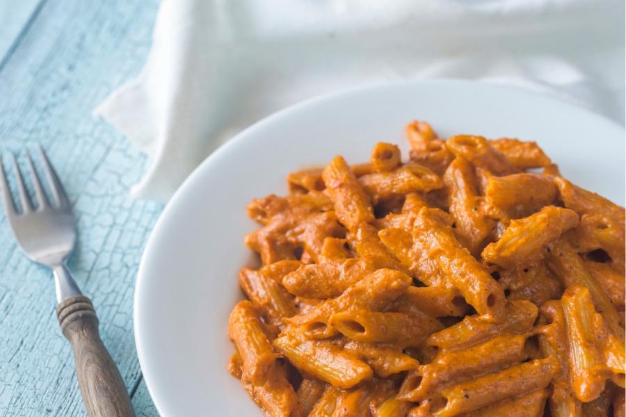 Penne con salsa cremosa de tomate (pasta a la vodka) en un plato blanco, con un tenedor sobre una mesa de madera azul clara.