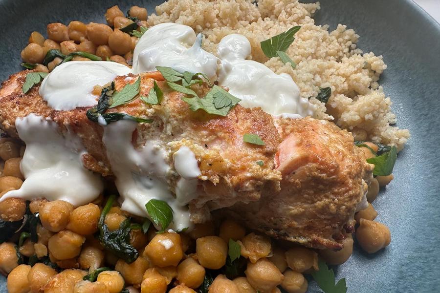 Lomo de salmón especiado y dorado, napado con salsa de yogur y espolvoreado de perejil, sobre garbanzos salteados con espinacas y con cuscús al lado, en un plato azul.