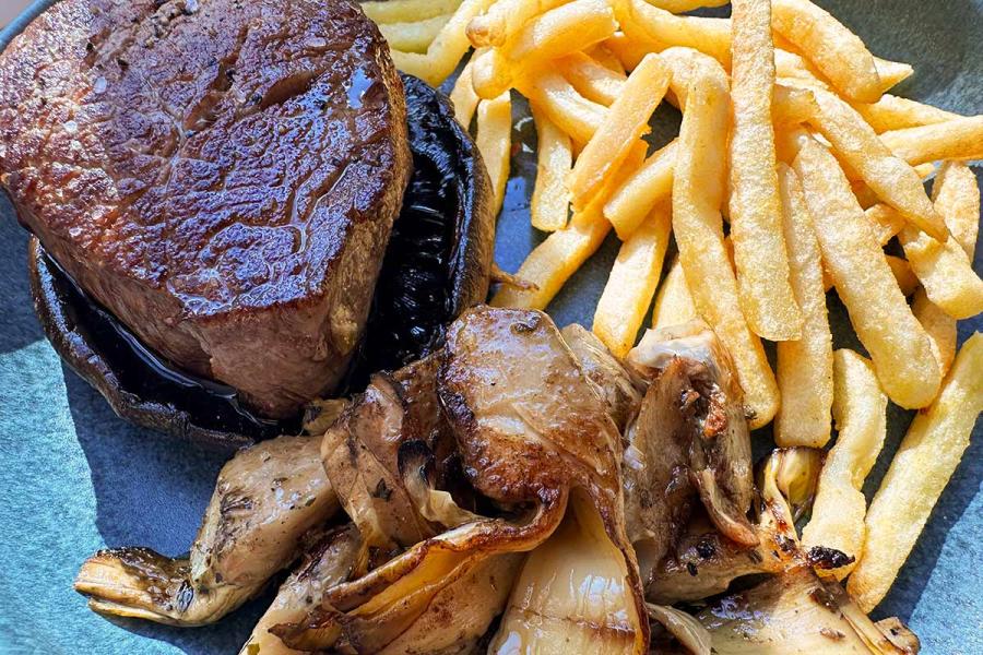 Solomillo de ternera a la plancha sobre un sombrero de portobello, acompañado de setas y alcachofas salteadas y de patatas fritas, en un plato azul.