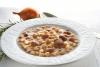 Un plato blanco de loza con pasta y alubias, una cebolla y una cuchara al fondo.