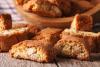 Unos biscotti de almendra sueltos sobre una mesa de madera y un recipiente de madera con más biscotti al fondo.