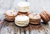 Dos macarons de vanilla y cuatro macarons de chocolate sobre una mesa de madera, con virutas de chocolate espolvoreadas alrededor.