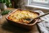 Un gratinado de lentejas y verduras recién horneado, servido en una fuente rústica de cerámica. El puré de patata dorado que lo cubre parece crujiente y ligeramente dorado. Se ve una rica mezcla de lentejas con zanahorias, cebolla, pimiento y una base de tomate.