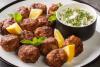 Albóndigas griegas fritas conocidas como keftedes, hechas con carne, servidas con tzatziki y trozos de limón en un plato de loza blanco.