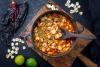 Un cuenco de pozole rojo rodeado de algunos de los ingredientes que se usan para hacerlo.