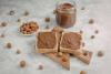 Dos tostadas untadas con pasta de chocolate y avellana con un tarro de vidrio lleno de pasta detrás. Están rodeados de avellanas peladas.