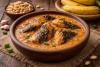 Cuenco de barro con salsa de cacahuete espesa y trozos de pescado ahumado, sobre una mesa de madera rústica, con cacahuetes, ajos y plátanos al fondo.