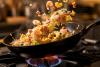Sartén salteando arroz tres delicias con gambas, guisantes, jamón, huevo y zanahoria sobre una llama de gas.