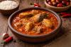 Cuenco de barro con salsa de palmiste rojiza y espesa sobre una mesa de madera rústica; alrededor hay arroz blanco, ajos, guindillas rojas y una cesta de frutos al fondo.