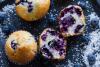 Muffins de arándanos azules sobre una superficie oscura, uno de ellos partido por la mitad mostrando su interior esponjoso con abundantes arándanos, rodeados de frutos frescos y coco rallado.