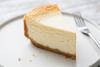Porción de tarta de queso clásica con base de galleta servida en un plato blanco, con un tenedor desenfocado al fondo.