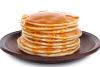 Torre de tortitas americanas doradas cubiertas con sirope de arce, servidas en un plato de cerámica oscura sobre un fondo blanco.