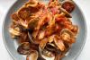 Plato hondo gris con cintas de pasta (tagliatelle) en salsa de tomate marinera rodeadas de almejas abiertas con sus valvas, sobre una mesa blanca.