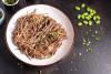 Cuenco blanco con fideos soba fríos aliñados, espolvoreados con cebolleta picada y hebras de alga, junto a granos de edamame y sésamo esparcidos sobre una superficie oscura.