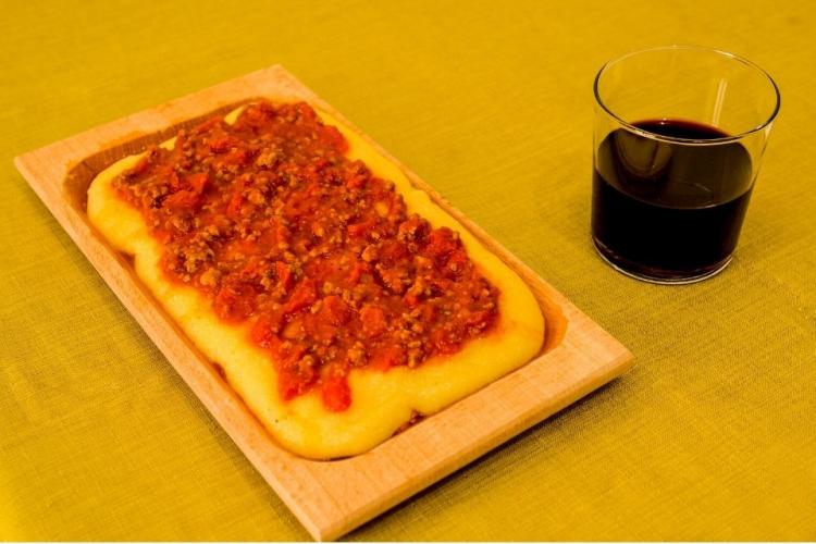 Una fuente de madera con polenta con salsa de carne y un vaso de vino tinto.