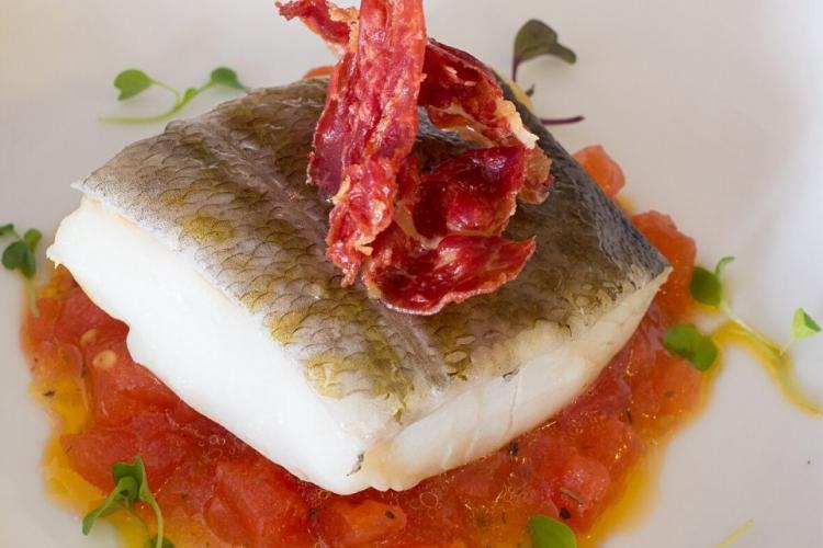 Trozo de pescado a la plancha sobre salsa de tomate adornado con jamón serrano.