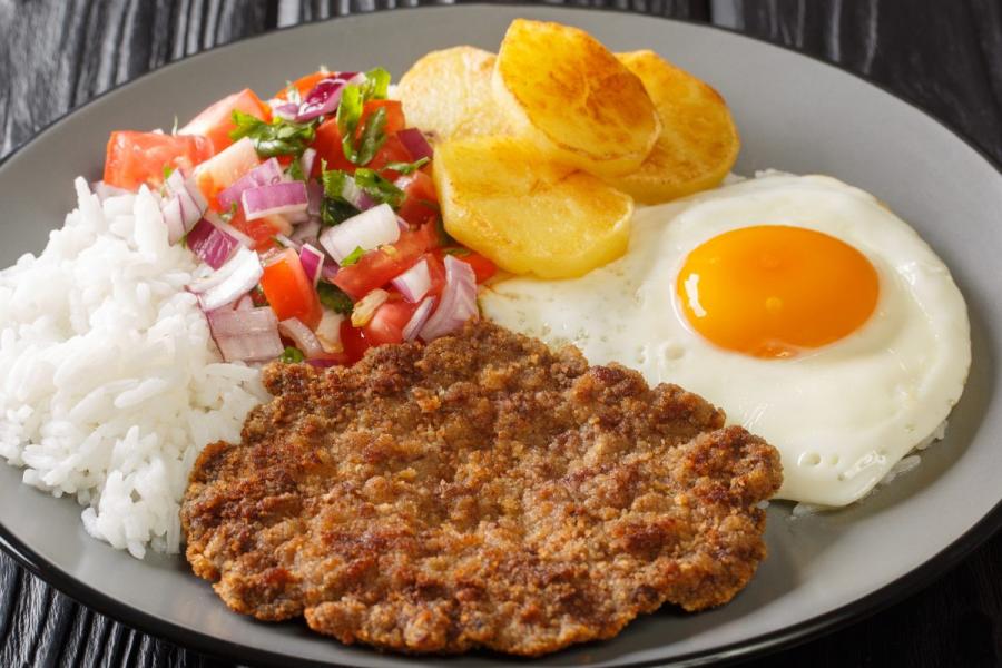 Silpancho, con filete de carne picaa empanado, huevo frito, patata en rodajas, arroz blanco y ensalada fresca de tomate y cebolla.