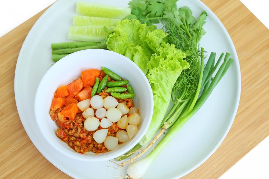Un cuenco blanco de salsa de chile nam prik ong adornado con cebollas en vinagre, zanahorias y chiles verdes, servido con lechuga fresca, pepino y cebollas verdes en un plato blanco.