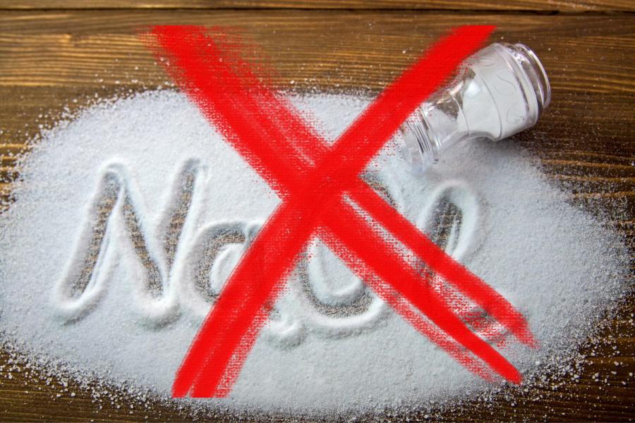 Un fondo de madera con sal esparcida formando la fórmula NaCl, con una gran X roja sobre la sal y un salero volcado al lado, indicando la prohibición del consumo de sal.