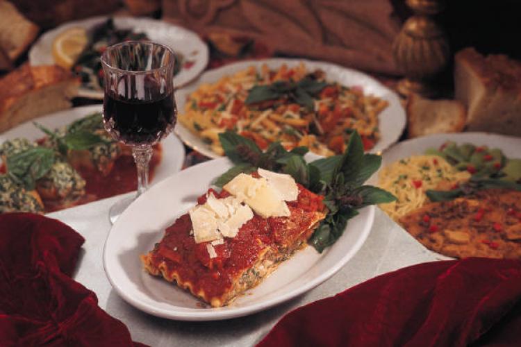 Mesa italiana con lasaña, otros platos de pasta, y una copa de vino tinto.